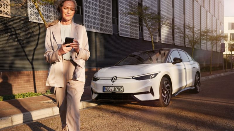 Eine Frau im Anzug schaut auf ihr Smartphone, im Hintergrund parkt ein weißer VW ID.7 am Straßenrand.