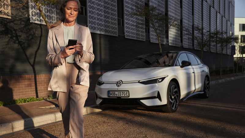 A woman in a suit looks at her smartphone, a white VW ID.7 is parked on the side of the road in the background.