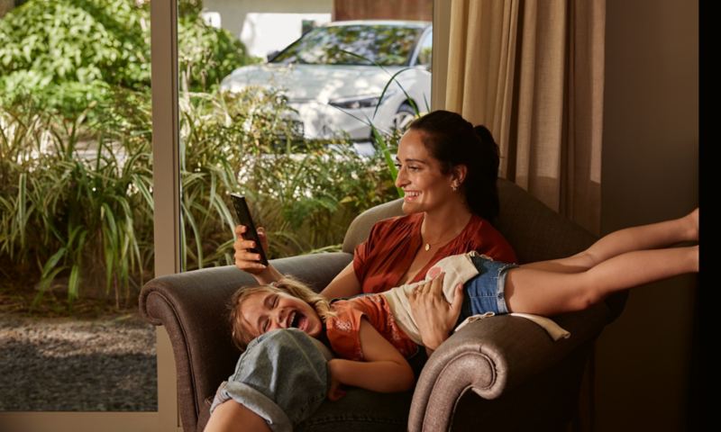 Une femme est assise sur un fauteuil avec un enfant souriant sur les jambes. Dans sa main droite, elle tient un smartphone. La VW ID.7 se trouve à l'extérieur, en arrière-plan.