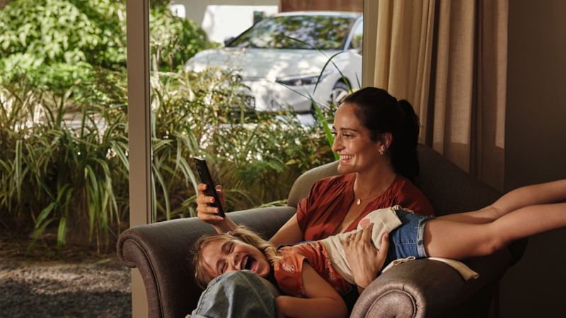 Alt Text: Mutter und Tochter sitzen auf einem Sessel und schauen sich den Volkswagen Connect Shop auf einem Smartphone an