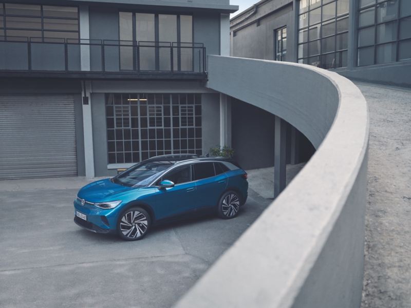 Una Volkswagen ID.4 blu è parcheggiata a lato di un complesso residenziale grigio di stile industriale e moderno.