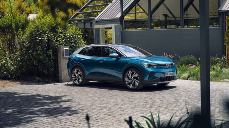 Une Volkswagen ID.4 bleue est garée de côté devant une maison moderne grise, et se recharge sur l’ID. Charger Wallbox. En arrière-plan, beaucoup de végétation.