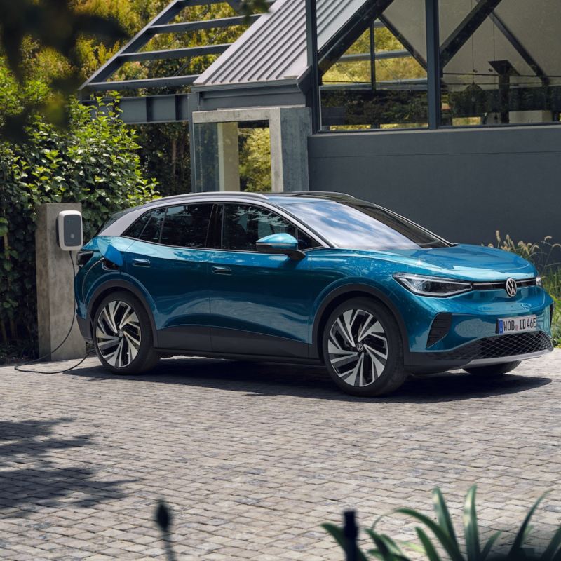 Ein blauer VW ID.4 parkt seitlich vor einem grauen, modernen Haus und lädt an der ID. Charger Wallbox. Im Hintergrund viel grün.