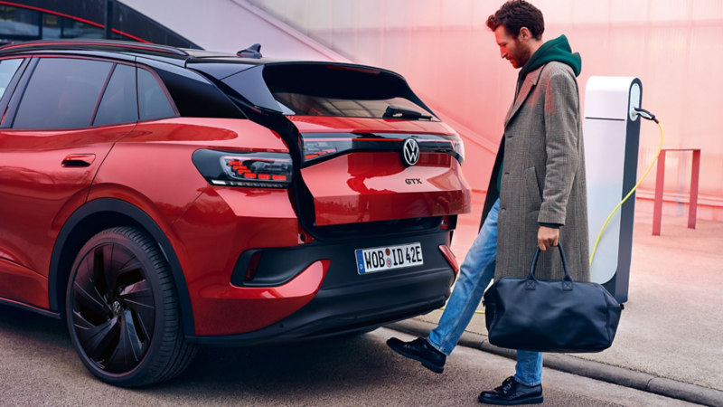 Ein Mann öffnet per Fußgeste den Kofferraum eines roten VW ID.4 GTX an einer Ladesäule.