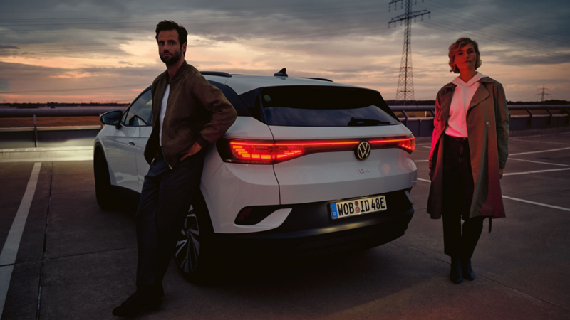 Ein weißer VW ID.4 steht auf einem Parkplatz in der Dämmerung. Die Heck-Lichtsignatur leuchtet ein Pärchen dahinter an.