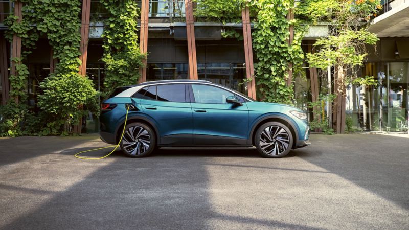 Vista laterale di una Volkswagen ID.4 blu collegata a un cavo di ricarica giallo. L’auto è parcheggiata di fronte a un edificio coperto di piante verdi.