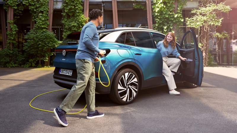 Ein Mann mit Ladekabel in der Hand geht zu einem blauen VW ID.4, der in einem begrünten Hof parkt.