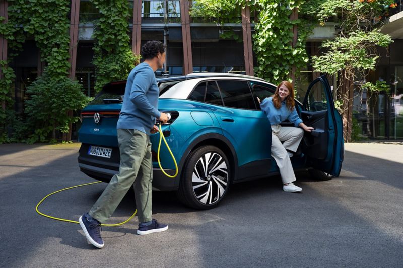 Ένας άνδρας με ένα κίτρινο καλώδιο φόρτισης για ηλεκτρικά αυτοκίνητα πλησιάζει το πίσω μέρος ενός σταθμευμένου μπλε VW ID.4. Μία νέα γυναίκα αποβιβάζεται από την πόρτα του συνοδηγού.