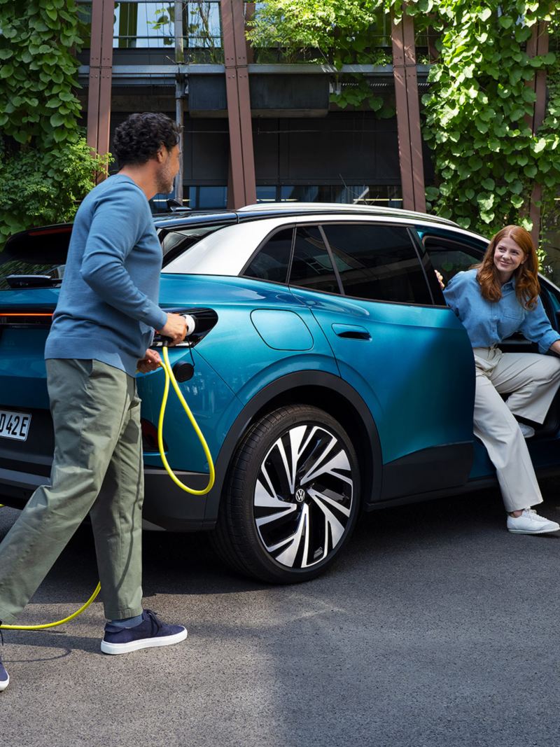 Un homme muni d’un câble de recharge jaune pour voitures électriques s’approche de l’arrière d’une Volkswagen ID.4 bleue stationnée. Une jeune femme descend de la porte passager.
