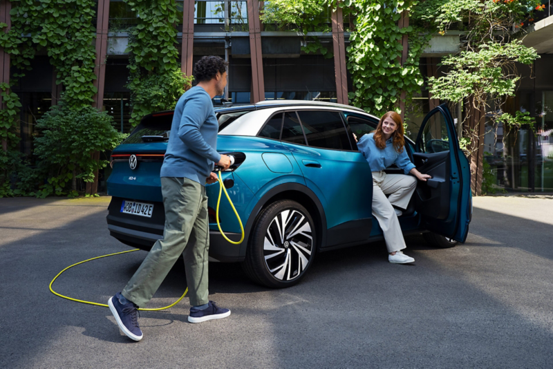 Ein Mann mit einem gelben Ladekabel für Elektroautos geht auf das Heck eines parkenden blauen VW ID.4 zu. Aus der Beifahrertür steigt eine junge Frau aus.