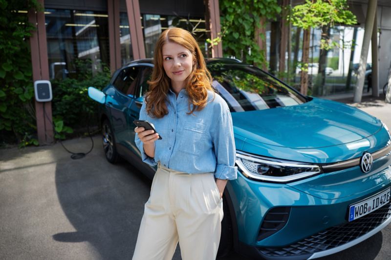 Frau mit Smartphone in der Hand steht vor einem blauen VW ID.4. Im Hintergrund ist eine Wallbox zu sehen, an die das Fahrzeug angeschlossen ist.