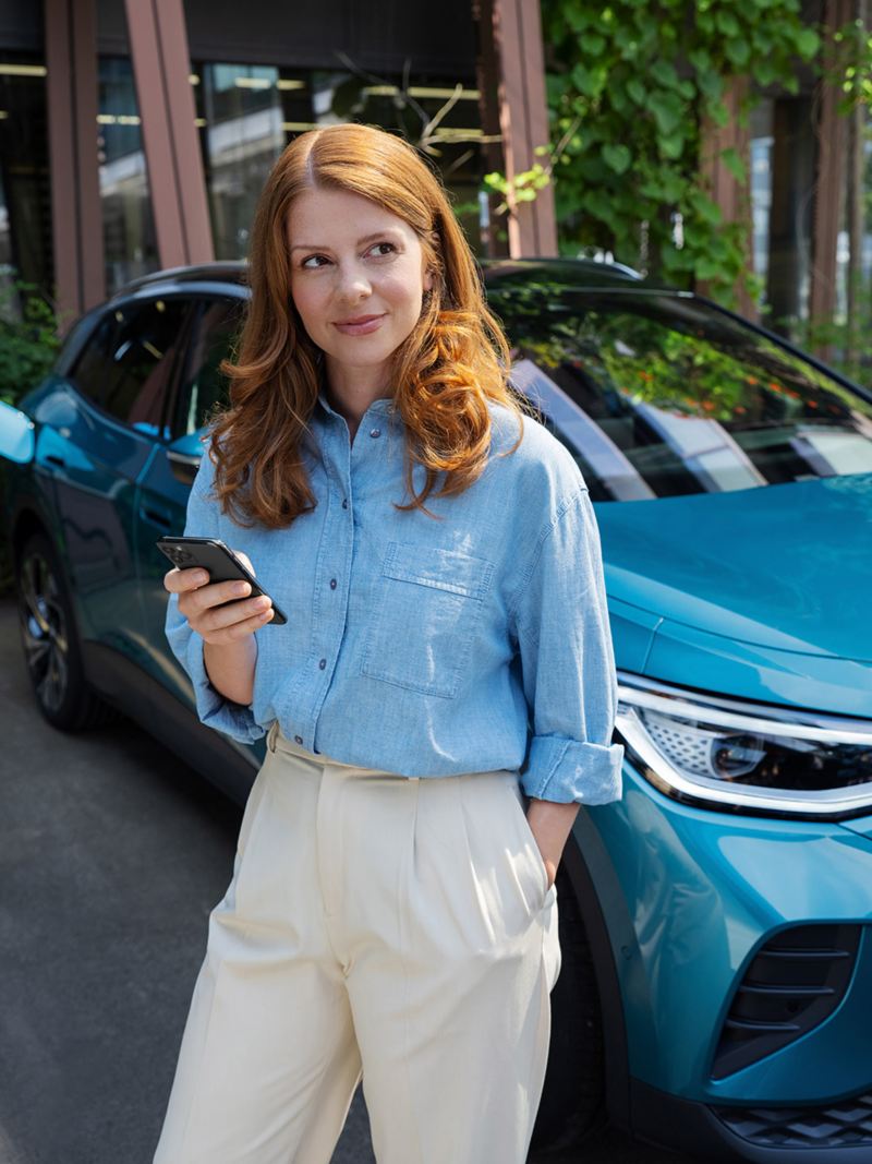Une femme tenant un smartphone à la main se tient devant une Volkswagen ID.4 bleue. En arrière-plan, on voit une Wallbox à laquelle le véhicule est connecté.