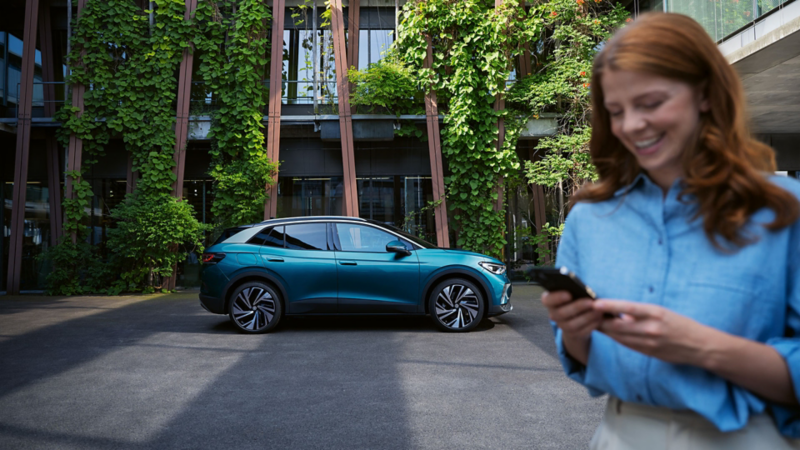 Junge Frau steht im Hof vor einem VW ID.4 Pro. Sie hält ein Handy in der Hand.