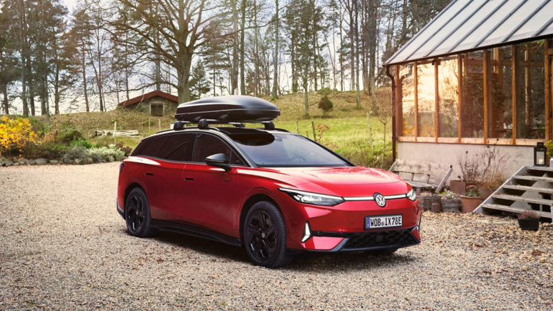Roter VW ID.7 GTX Tourer mit Dachbox parkt zwischen zwei Holzhäusern, im Hintergrund ein Wald.
