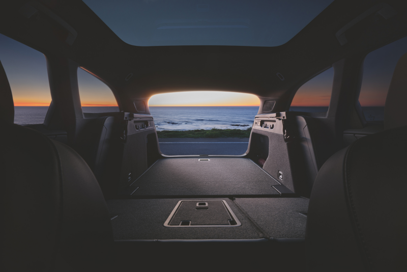 Interier shot van een Volkswagen ID.7 Tourer waarbij de achterbank is neergeklapt en de kofferbak open staat, op de achtergrond kun je de zee zien met ondergaande zon