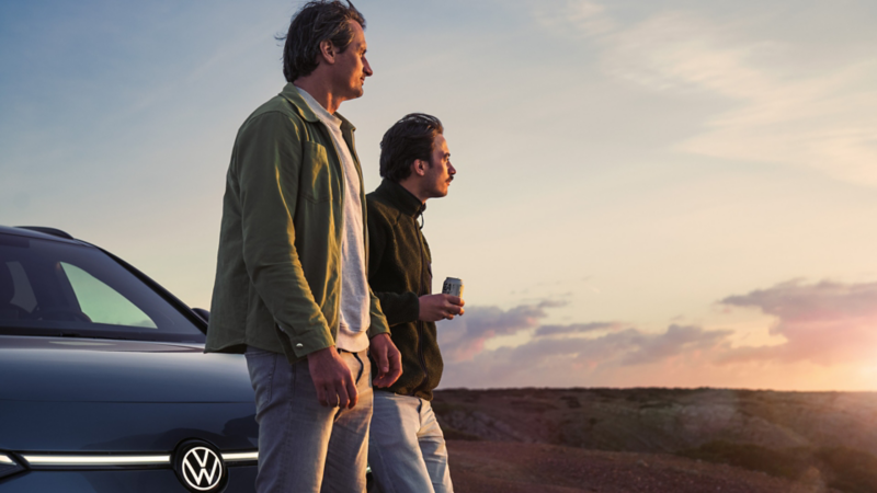 Zwei Männer lehnen an der Motorhaube eines VW ID.7 Tourer und schauen auf einen Sonnenuntergang am Meer.
