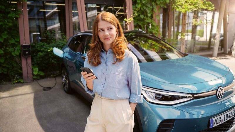 Frau mit Smartphone in der Hand steht vor der Motorhaube eines blauen VW ID.4, der an einer Wallbox angeschlossen ist.