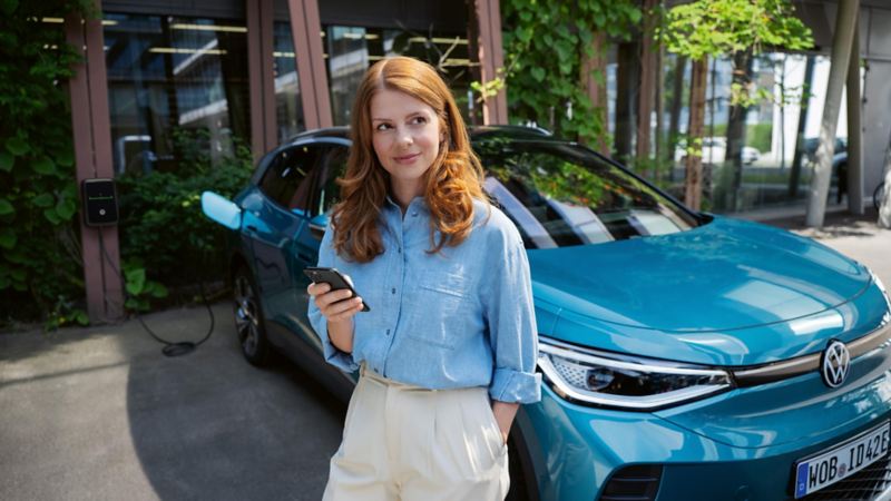 Frau mit Smartphone in der Hand steht vor einem blauen VW ID.4. Im Hintergrund ist eine Wallbox zu sehen, an die das Fahrzeug angeschlossen ist.