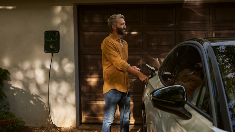 Ein Mann lädt einen weißen VW ID.7 Pro an einer Wallbox vor einem Haus.