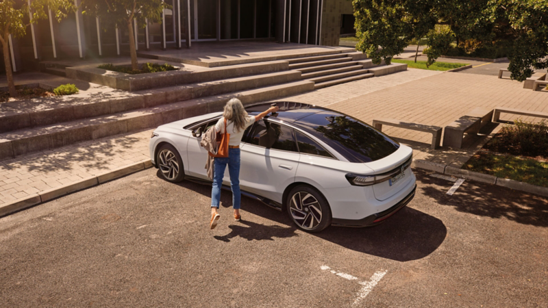 Frau nimmt einen Becher vom Dach eines VW ID.7, der vor einem Bürogebäude parkt.