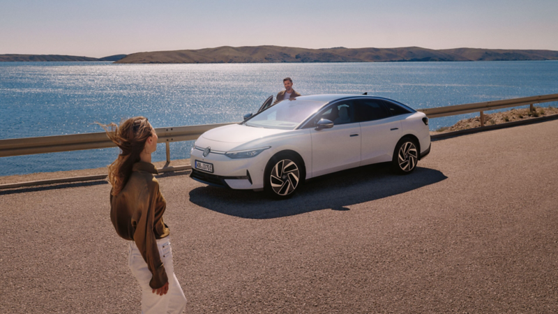 Eine Frau geht auf einen weißen VW ID.7 zu, der am Meer parkt. Auf der Beifahrerseite steigt ein Mann aus.