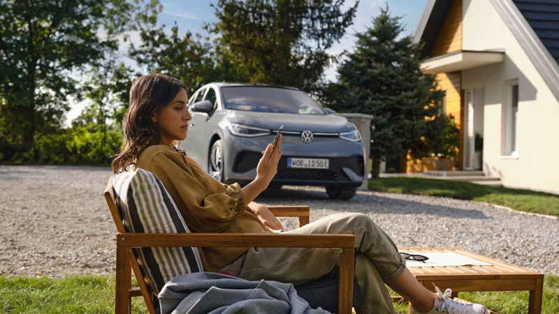 Frau sitzt entspannt im Gartenstuhl vor einem Haus und schaut auf ihr Smartphone; im Hintergrund steht ein Volkswagen ID. 