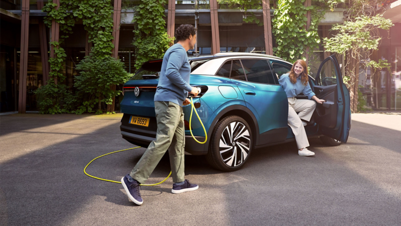 Une femme sort d'une ID.4 bleue côté passager. Un homme est en train de brancher un câble pour recharger l'ID.4.