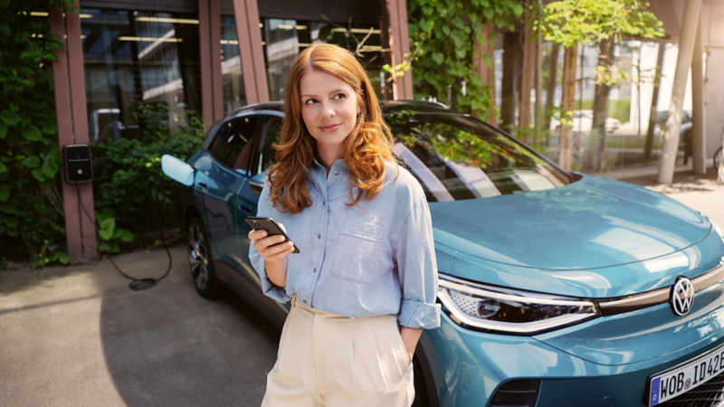 Frau steht mit Smartphone vor einem  Volkswagen ID. an einer Ladestation.