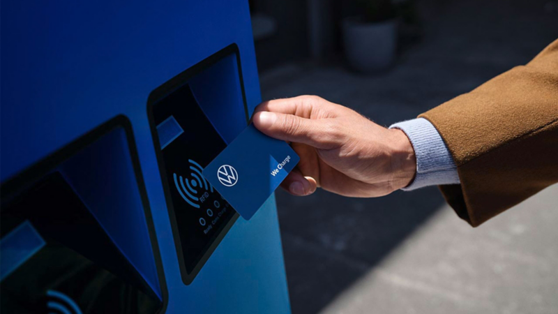 Eine Hand hält eine VW We Charge Ladekarte an eine blaue Ladestation.