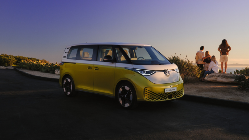 Un VW ID. Buzz giallo e bianco si trova da un lato sul bordo di una passeggiata sulla spiaggia. Sullo sfondo una famiglia con due bambini guarda il tramonto sul mare.