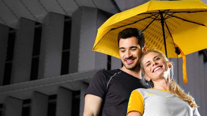Un ragazzo e una ragazza sorridenti sotto un ombrello giallo originale Volkswagen.