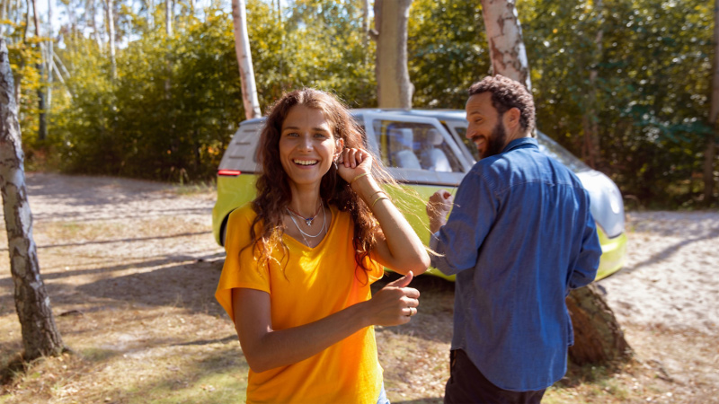 Smiling woman in an orange shirt and a man in a blue shirt standing near a green and white van in a sunny, wooded area.