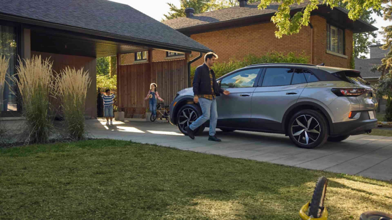 Un homme en veste se dirige vers un VUS électrique Volkswagen ID.4 argenté stationné dans une allée résidentielle. Deux enfants jouent dans l'abri de voiture attenant à une maison moderne, avec une maison en brique et des arbres visibles en arrière-plan sous un soleil chaleureux.