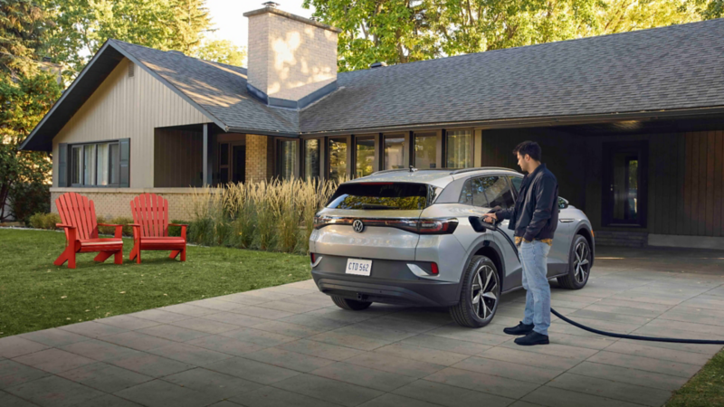 : A person plugging a charging cable into the rear charging port of a Volkswagen ID.4 electric SUV parked in a residential driveway, with a house visible in the background.