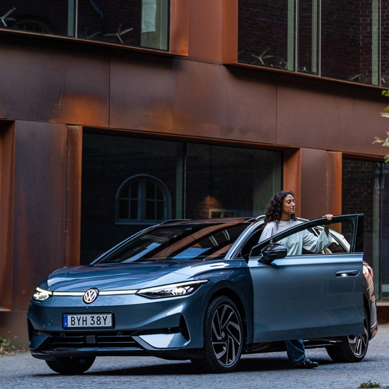 En ljusblå VW ID.7 Tourer parkerad i stadsmiljö. En kvinna stiger ur bilen.