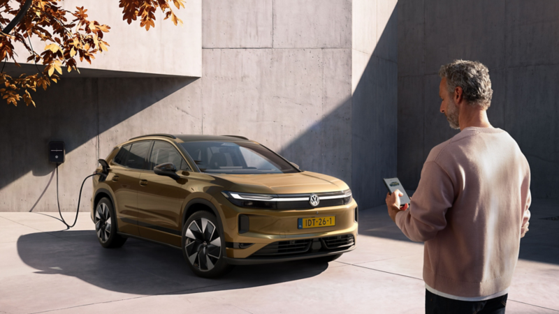 Een man kijkt op zijn telefoon. Op de achtergrond laadt een olijfbronskleurige VW ID. Tiguan op aan de laadpaal thuis.   