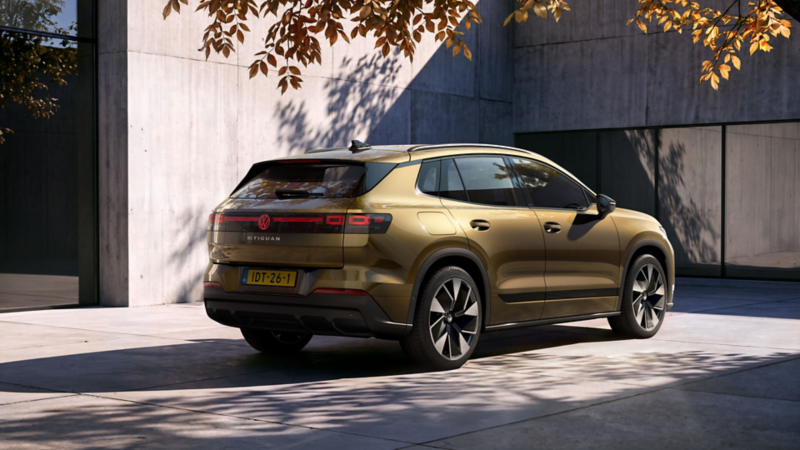 De achterkant en zijkant van een olijfbronskleurige VW ID. Tiguan. De auto staat geparkeerd voor een modern gebouw.   