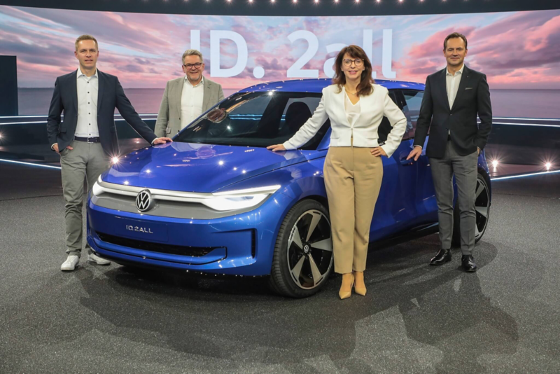 Λήψη ενός VW ID.2 all concept σε μπλέ χρώμα σε μια εκθεσιακή αίθουσα, με 4 ανθρώπους γύρω του.