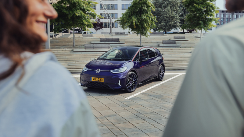 Image montrant l'avant d'une Volkswagen ID.3 GTX violette, garée sur un parking. Deux personnes, floues au premier plan, regardent vers la voiture.