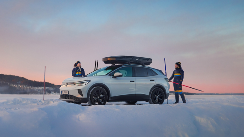En Volkswagen ID.4 står parkerad i vinterlandskap med en takbox på taket. Två skidåkare i landslagskläder går bredvid bilen