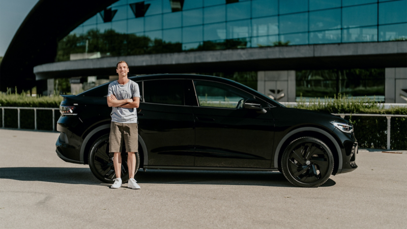 Un homme souriant, portant un t-shirt et un short, se tient les bras croisés à côté d'une Volkswagen ID.5 noire. La voiture est garée devant un bâtiment moderne aux grandes baies vitrées.