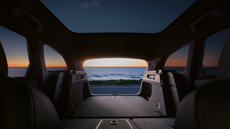  Vue du coffre d'une Volkswagen ID.7 Tourer, avec les sièges arrière rabattus pour créer un grand espace de chargement. Le soleil se couche sur l'océan, visible à travers le hayon ouvert.