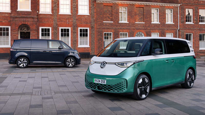 Two Volkswagen ID. Buzz vehicles parked on a paved urban street in front of a red brick building. The foreground features a green and white ID. Buzz, while a dark blue ID. Buzz is parked further back.