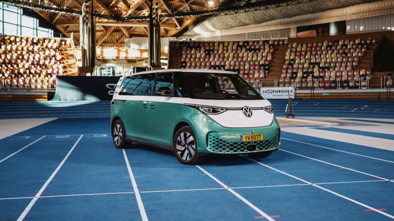 Volkswagen ID. Buzz bicolore vert et blanc stationné sur une piste d’athlétisme couverte à la Coque, avec des gradins multicolores en arrière-plan.