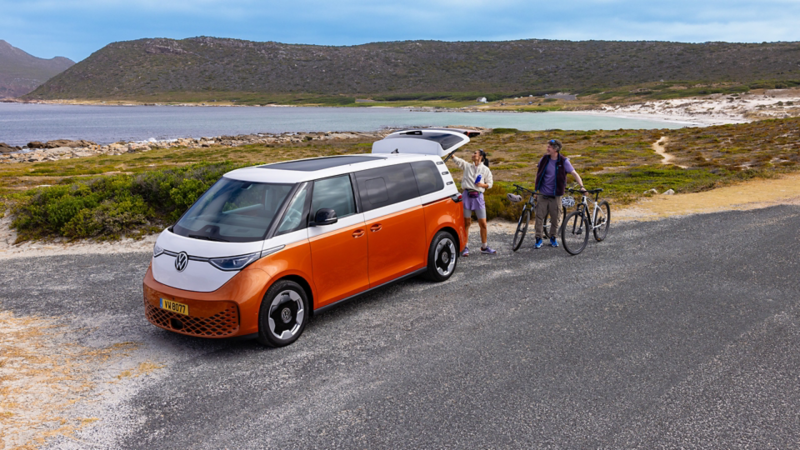 Van Volkswagen ID. Buzz Pro orange et blanc garé au bord de la mer, coffre ouvert, avec deux personnes et des vélos près de la plage.