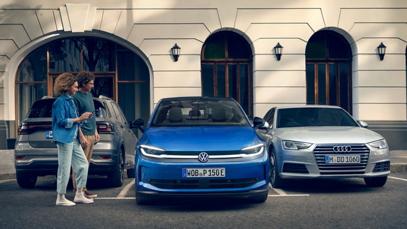 A woman holds her smartphone in her hand, next to her a blue VW ID. Polo is parking driverless backwards into a parking space.