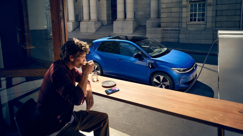 Ein Mann sitzt in einem Café, durch ein offenes Fenster ist der blaue VW ID. Polo zu sehen, der an einer Ladestation lädt.