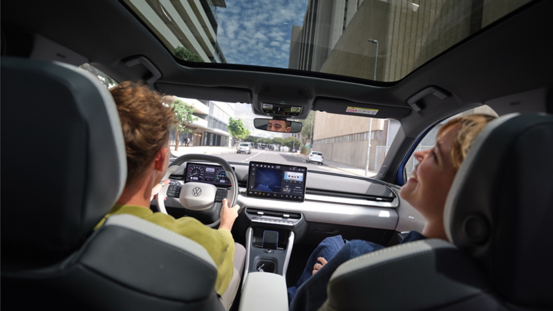 A man is driving a VW ID. Polo, with a laughing woman in the passenger seat. High-rise buildings can be seen through the panoramic roof.