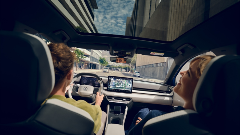 A man is driving a VW ID. Polo, with a laughing woman in the passenger seat. High-rise buildings can be seen through the panoramic roof.