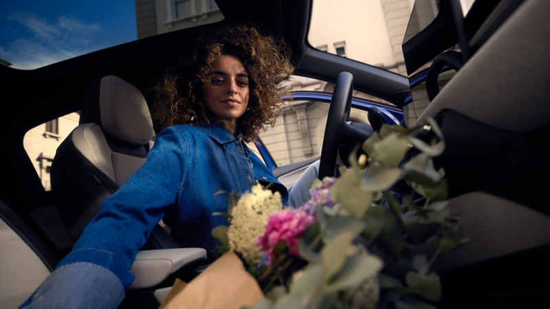 A woman is sitting in the driver's seat of a blue VW ID. Polo and reaches for a bouquet of flowers on the passenger seat.